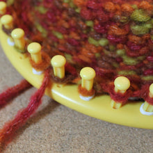 Load image into Gallery viewer, Close-up of a yellow knitting loom with multicolored yarn on a textured surface